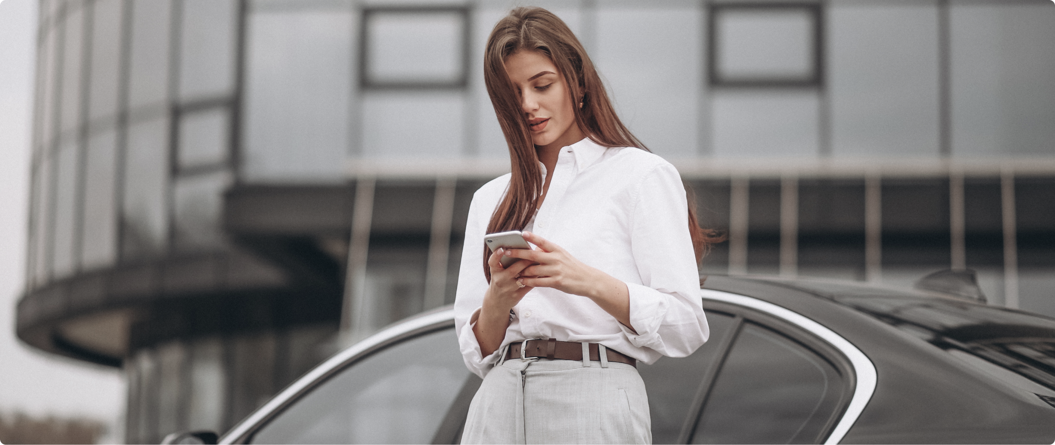 Mulher com cabelo castanho, vestindo camisa social branca para dentro da calça, segurando um celular e encostada em um carro.