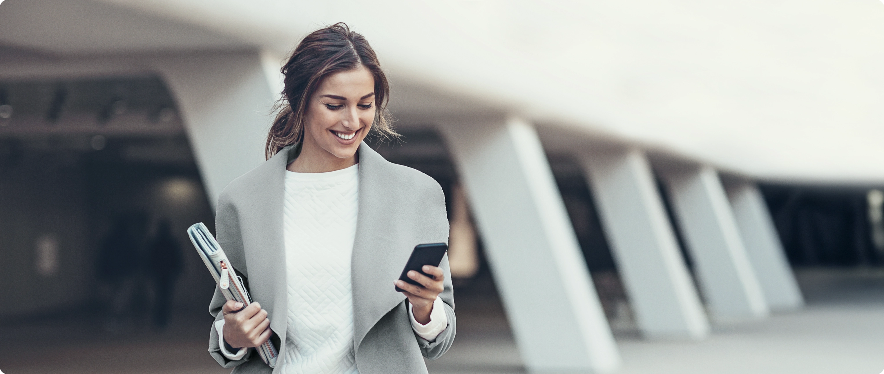 Mulher sorrindo enquanto caminha com celular e pasta em ambiente urbano.