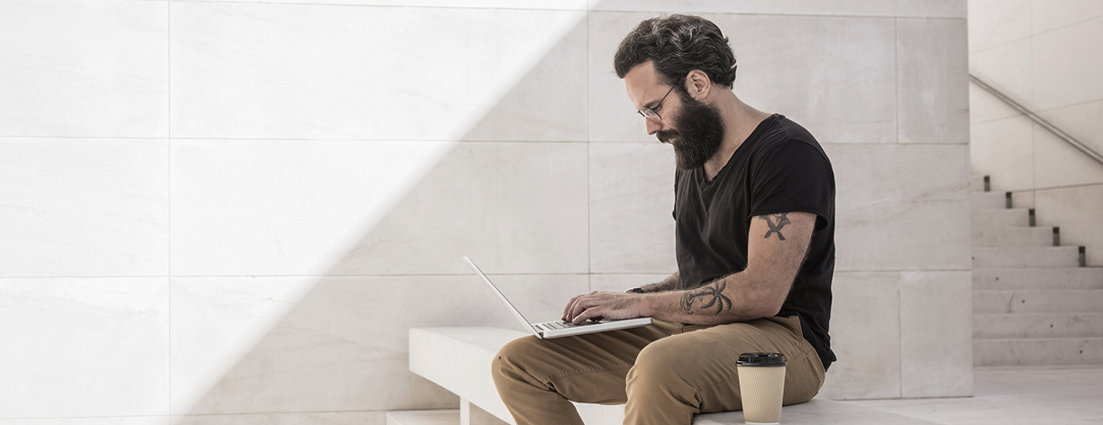 Homem tatuado usa notebook em ambiente claro com escada ao fundo.