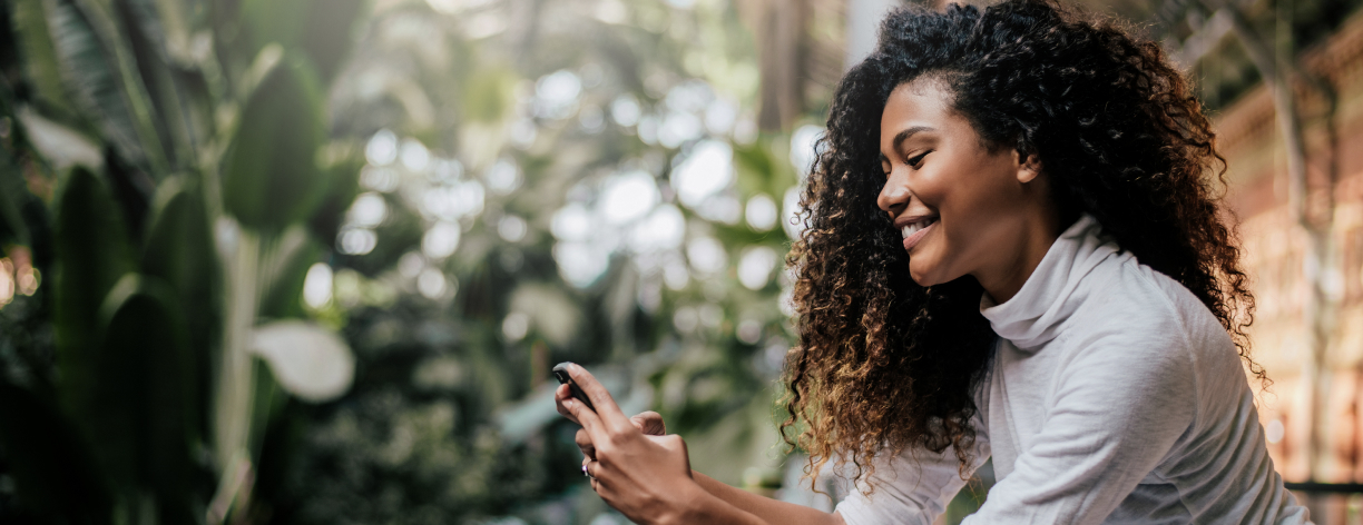 Mulher sorridente usa celular em local com vegetação.