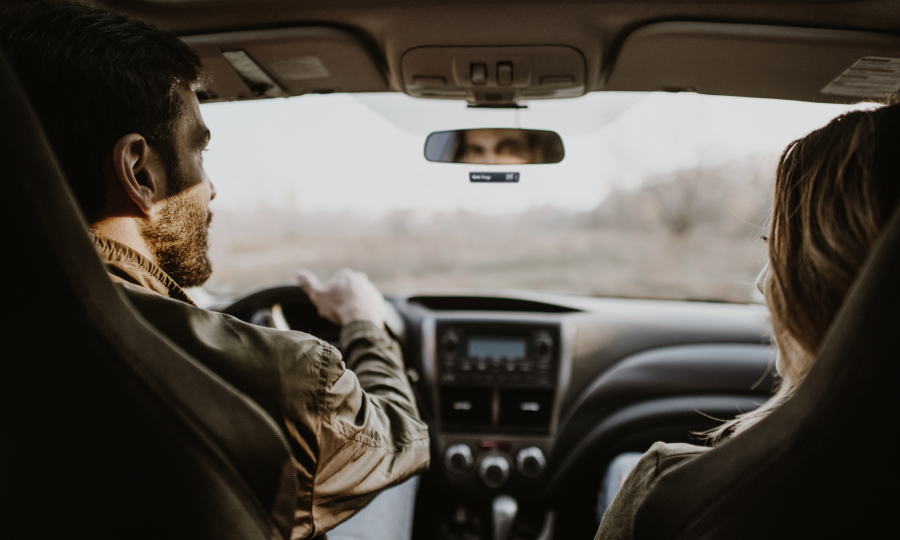 Homem dirigindo um carro, com uma mulher ao lado e paisagem ao fundo.