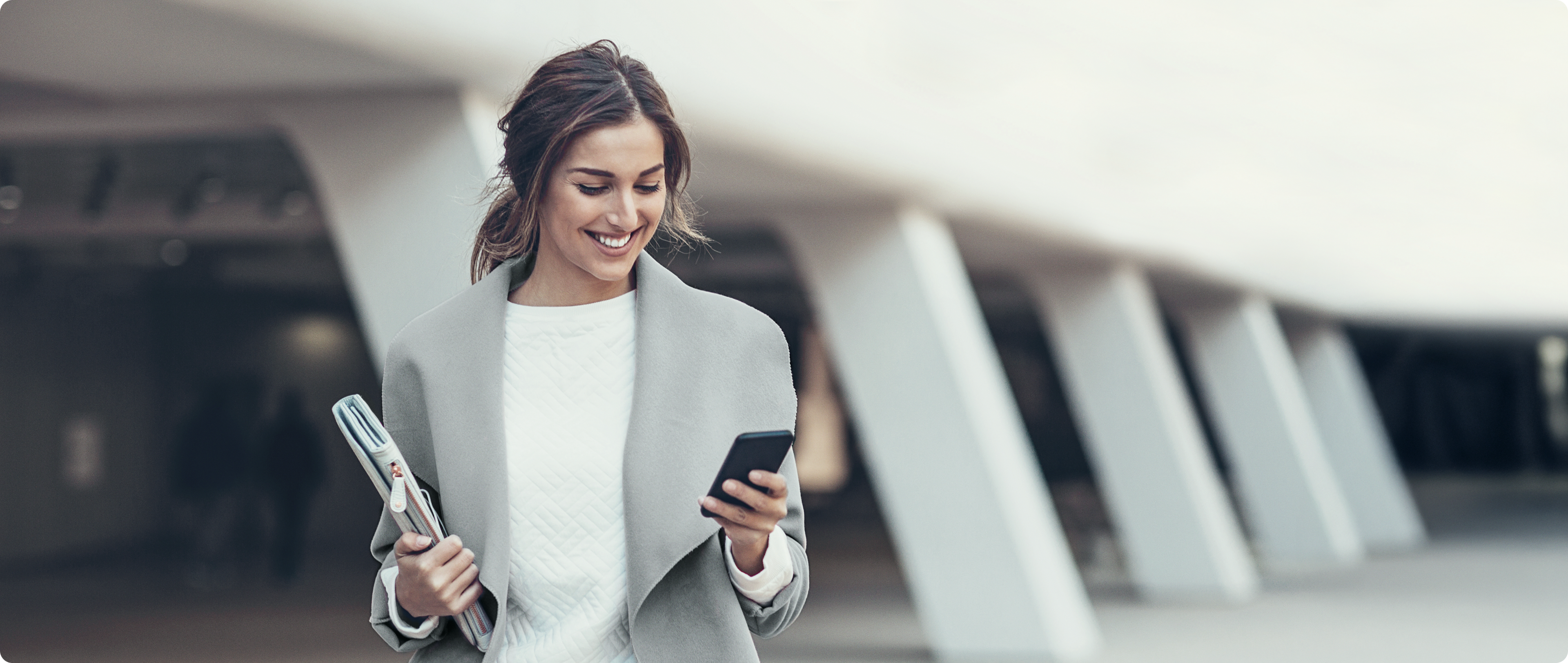 Mulher sorrindo enquanto caminha com celular e pasta em ambiente urbano.
