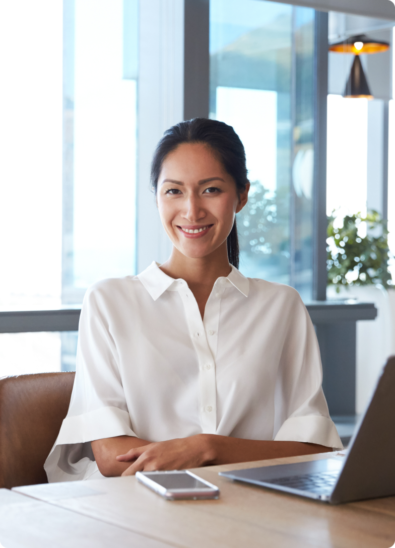 Mulher sorridente sentada à mesa com celular e notebook em ambiente de escritório.