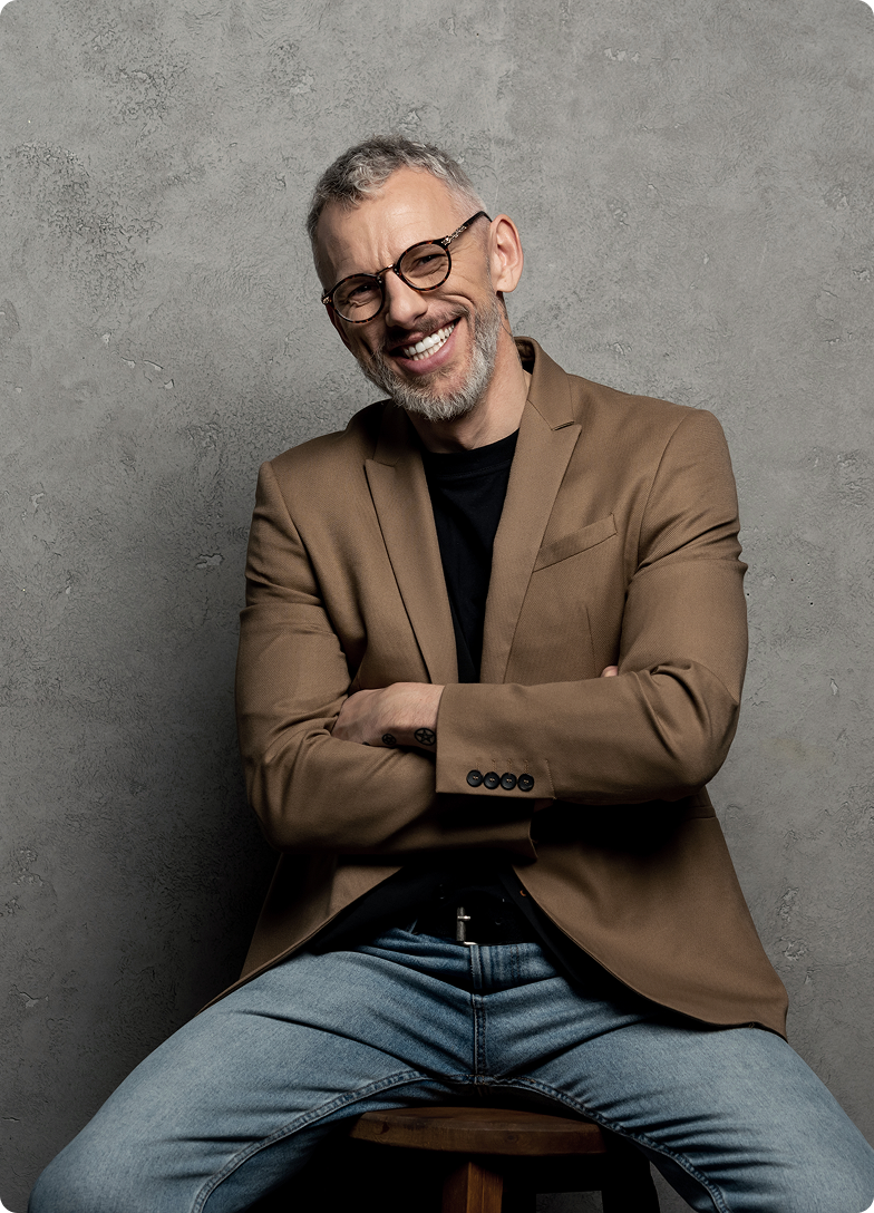 Homem grisalho sorridente, de blazer marrom e jeans, sentado com os braços cruzados.