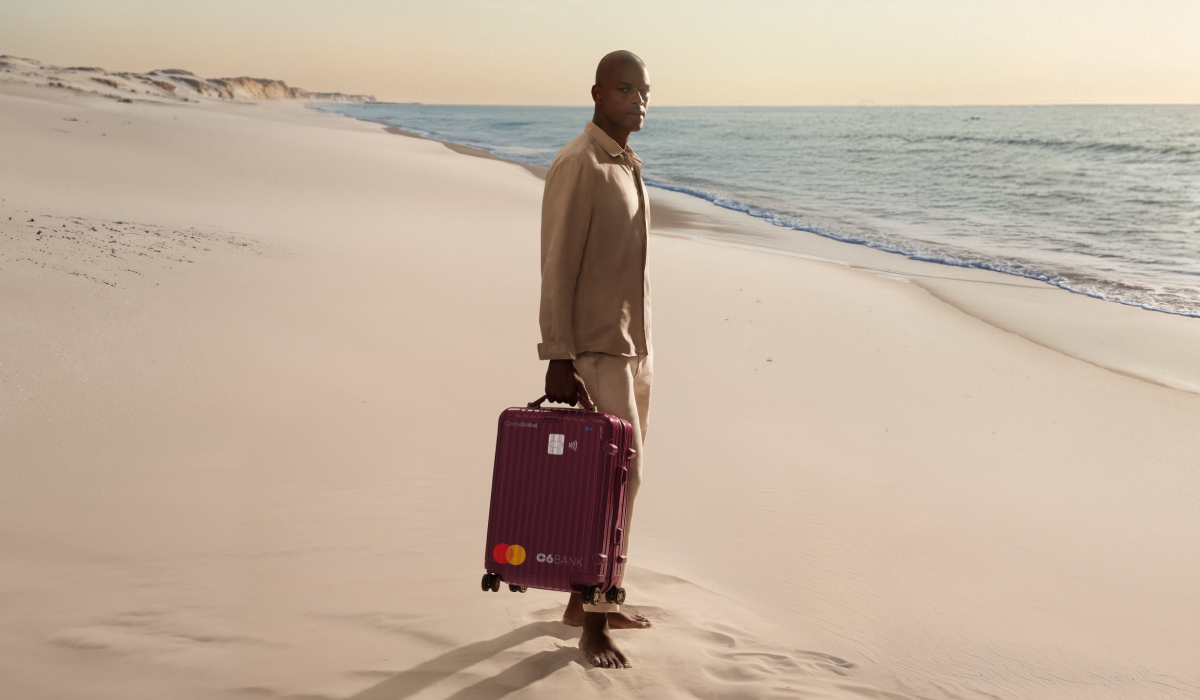 Homem na praia puxa mala com layout do C6 Conta Global. Cena ao entardecer transmite mobilidade e tranquilidade.