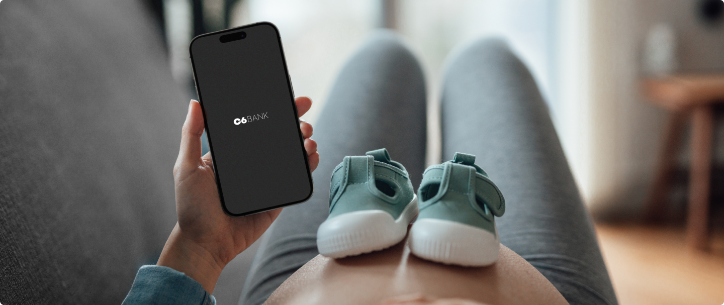Mulher grávida segura um celular C6 Bank com sapatinhos sobre a barriga.