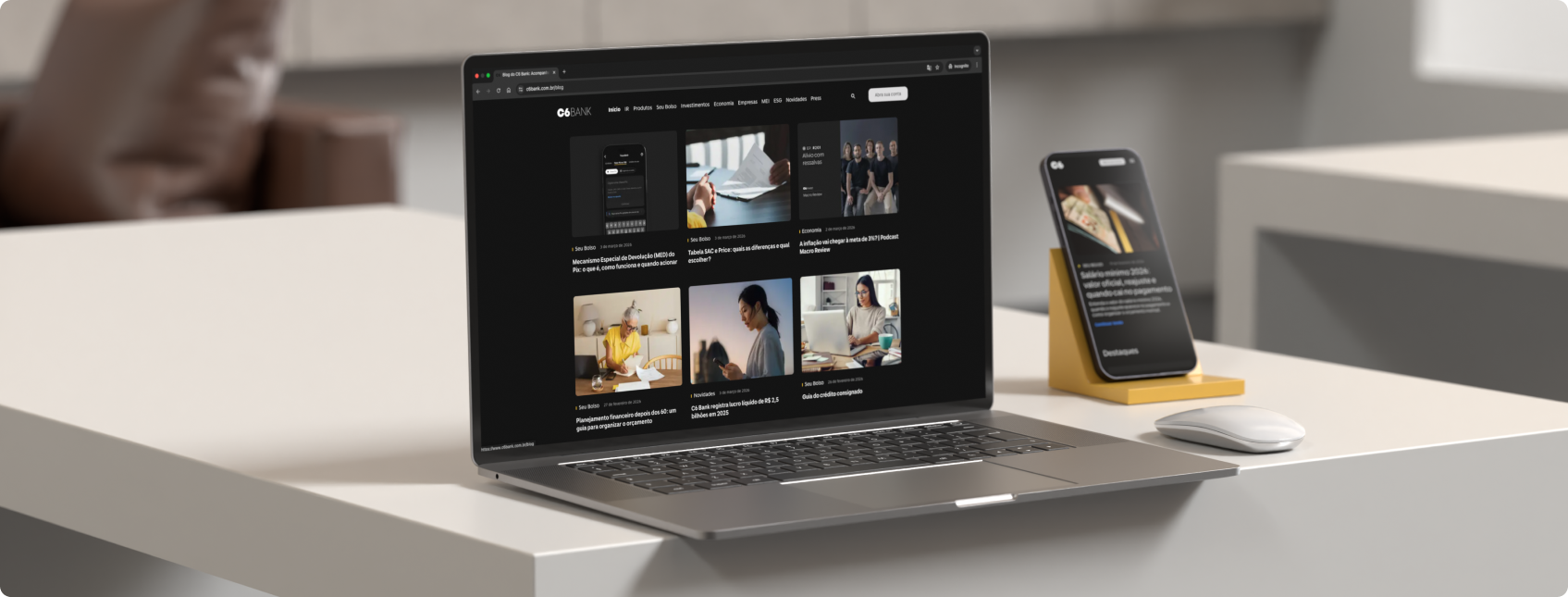 Notebook com blog do C6 Bank aberto ao lado de celular em suporte amarelo.