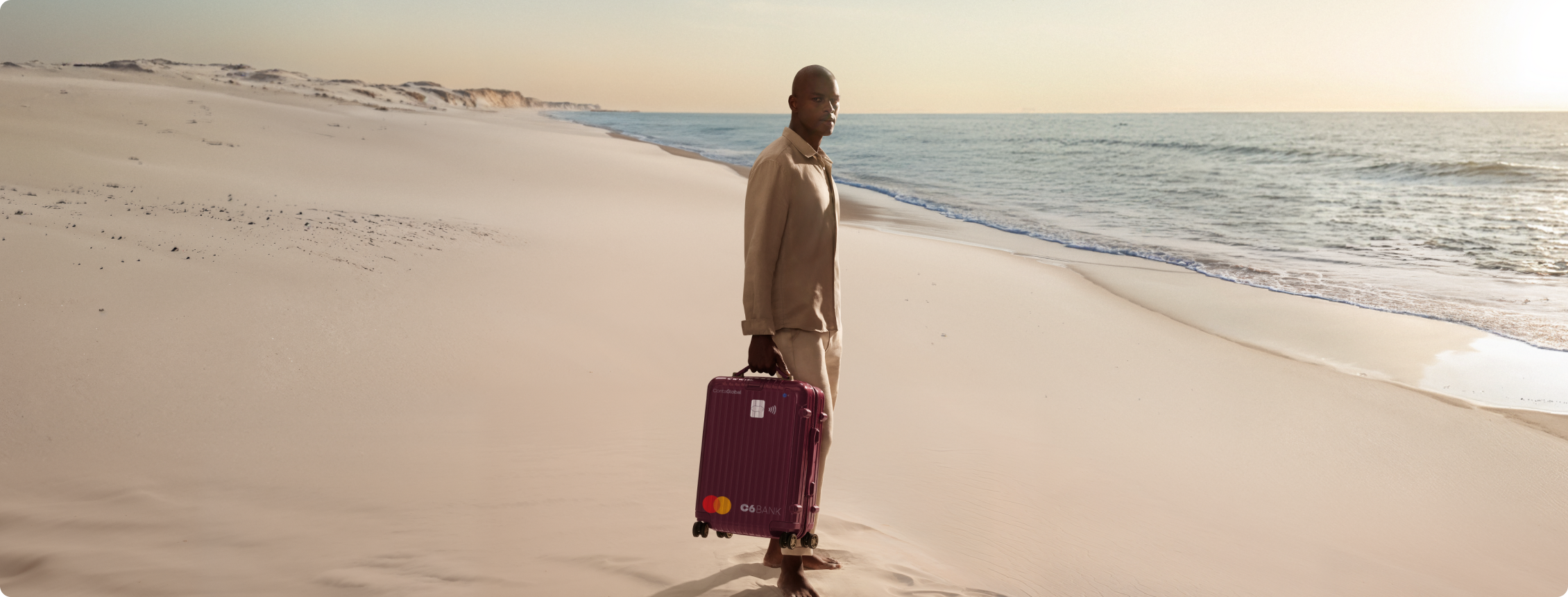 Homem na praia puxa mala com layout do C6 Conta Global. Cena ao entardecer transmite mobilidade e tranquilidade.