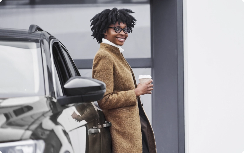 Mulher sorridente de óculos, com casaco marrom, encostada em um carro e segurando um café.