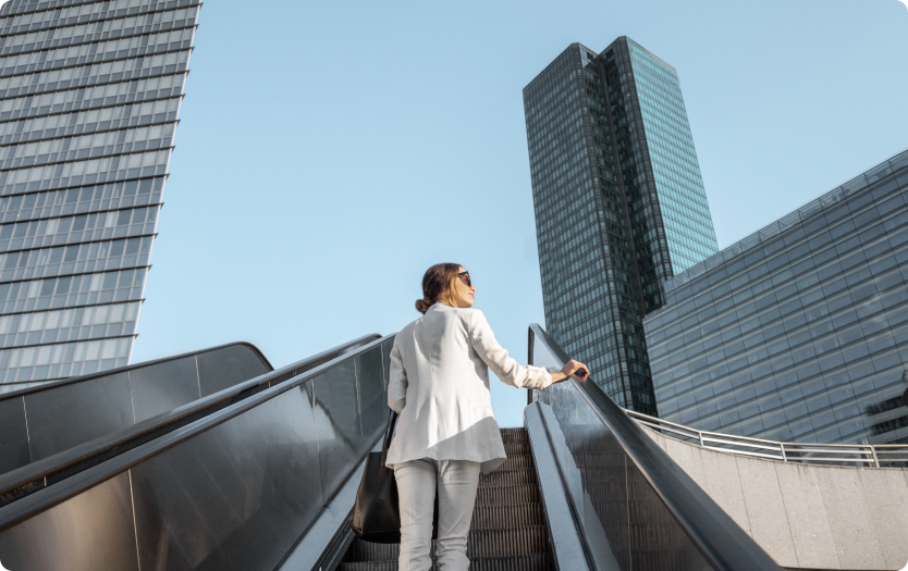 Mulher de terno branco sobe escada rolante entre prédios modernos.