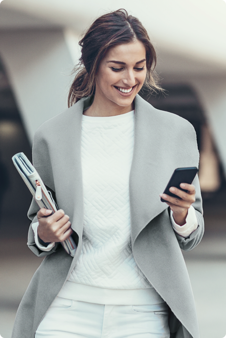 Mulher caminha olhando o celular e segurando cadernos em ambiente urbano.