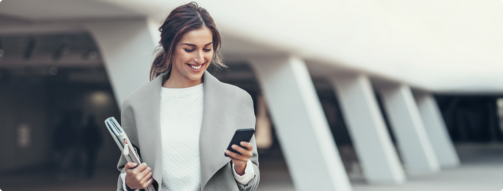 Mulher sorrindo enquanto caminha com celular e pasta em ambiente urbano.