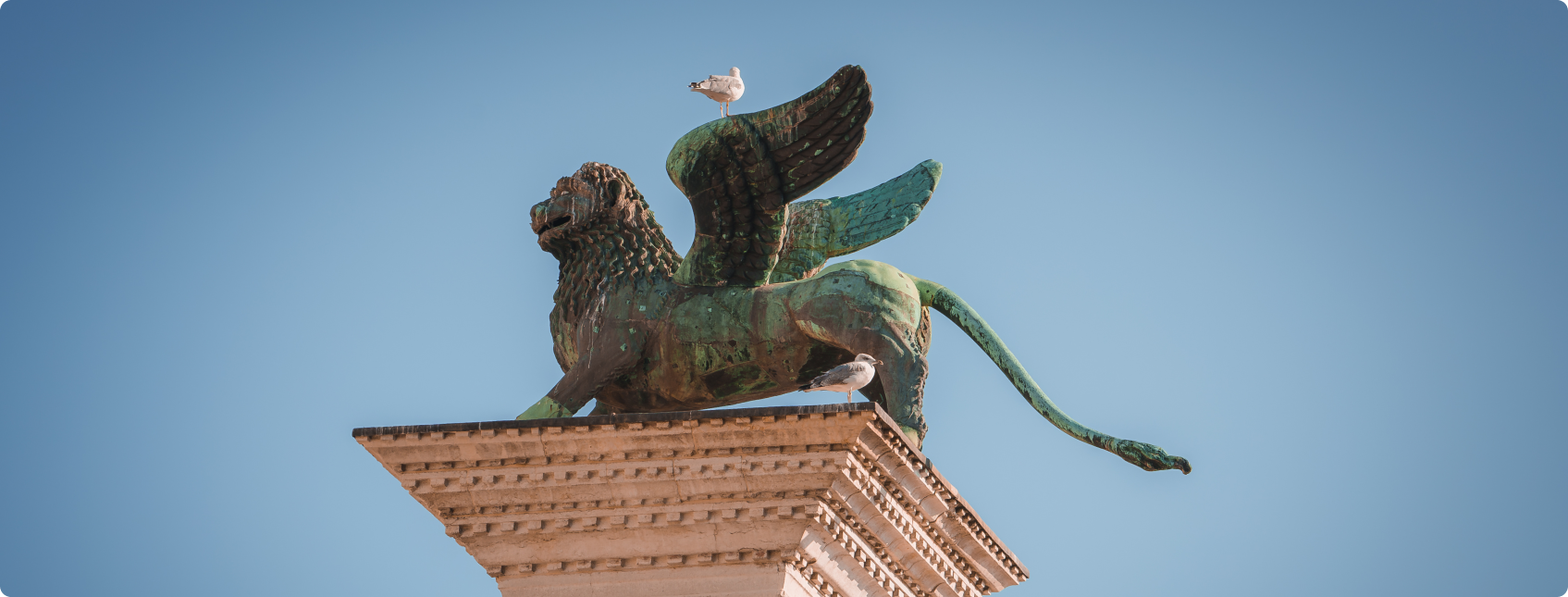 Leão de São Marcos alado de bronze com gaivotas pousadas em pedestal sob céu azul em Veneza.