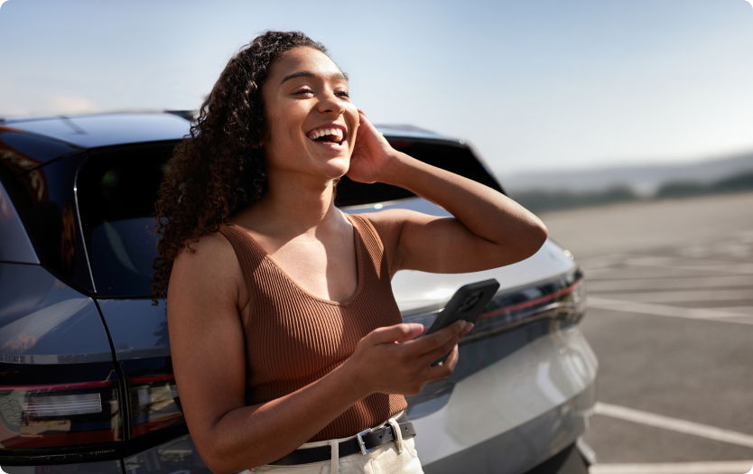 Mulher sorridente com celular ao lado de um carro em estacionamento aberto.