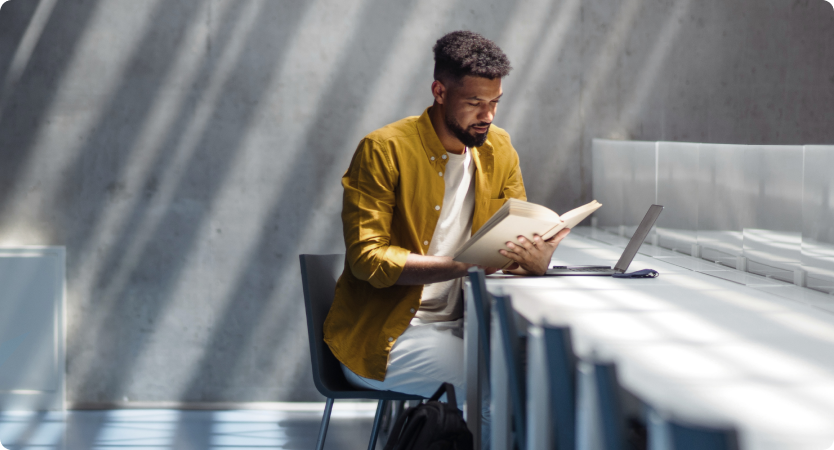 Homem com camisa mostarda lê livro ao lado de notebook em ambiente iluminado.