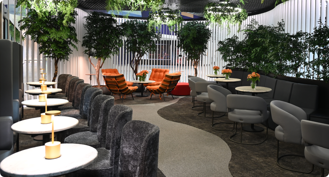 Espaço interno elegante com poltronas, mesas redondas, plantas e arranjos florais sobre as mesas.