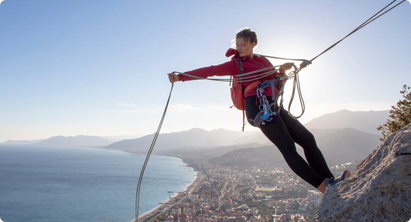 Mulher escalando montanha segura cordas com cidade e mar ao fundo.