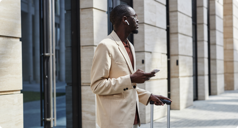 Homem elegante com celular e mala está parado em frente a prédio moderno.