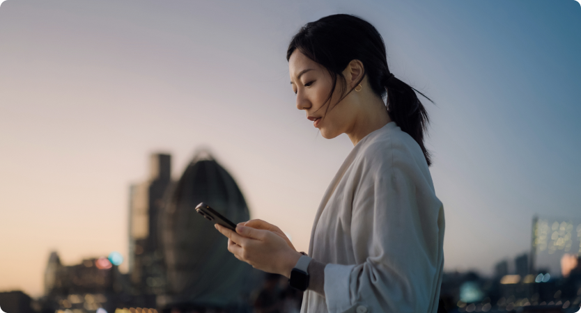 Mulher com celular em mãos observa a tela ao entardecer na cidade.