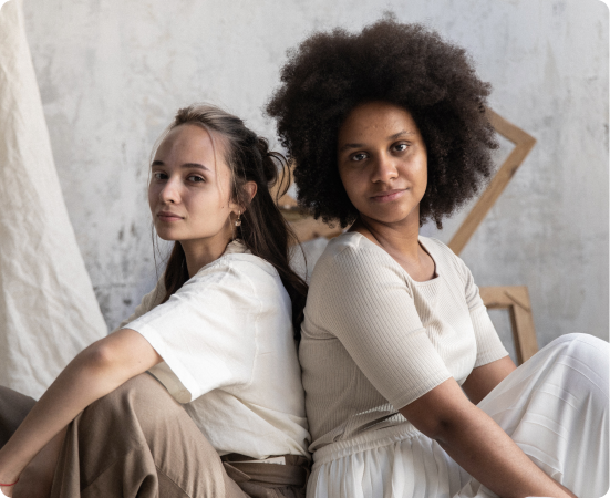 Fotografia de duas mulheres sentadas de costas uma para a outra, em um fundo cinza, com pano branco e molduras de madeira.