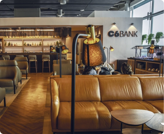 Lounge do C6 Bank com sofás caramelo, bar ao fundo e atmosfera sofisticada.