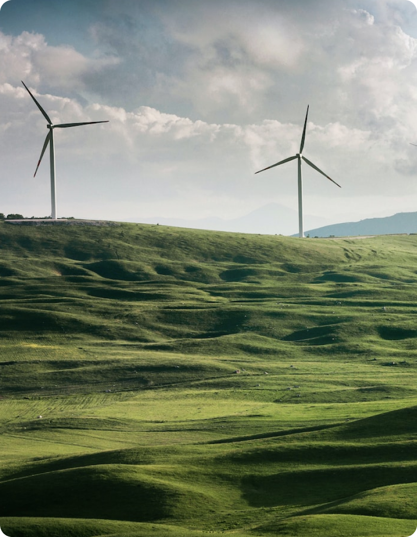 Fotografia de montanhas baixas, com dois moinhos de ventos e céu predominantemente nublado.