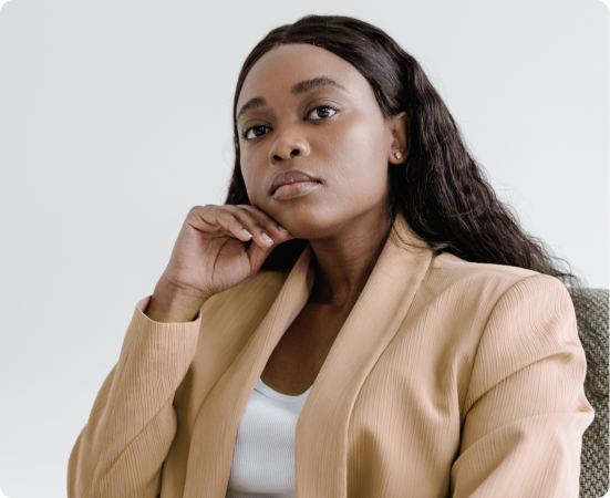 Fotografia de mulher negra com cabelo solto, de blazer bege e camiseta branca por embaixo, com a mão no queixo.
