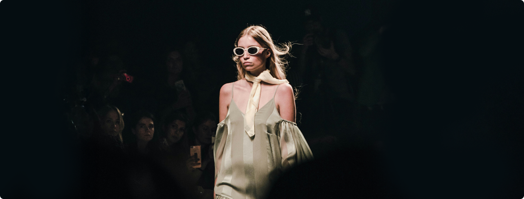 Modelo com vestido claro e óculos desfila em passarela com plateia ao fundo.