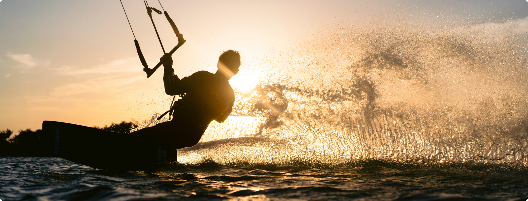 Kitesurfista em ação ao pôr do sol, com água respingando iluminada ao fundo.