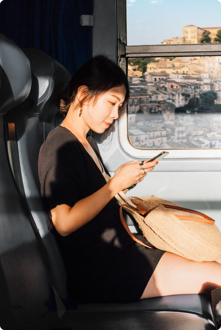 Mulher em trem usa o celular enquanto aprecia a vista de uma cidade histórica pela janela.