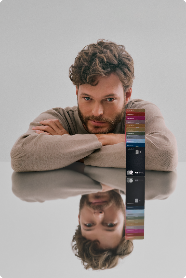 Foto de um homem branco de cabelos e barba castanhos claro, onde ele apoia seus braços em uma mesa refletiva com múltiplos cartões C6 Bank de diversas cores empilhados um em cima do outro ao seu lado. Ele veste um suéter bege, tem olho claro e tem uma expressão facial confiante.