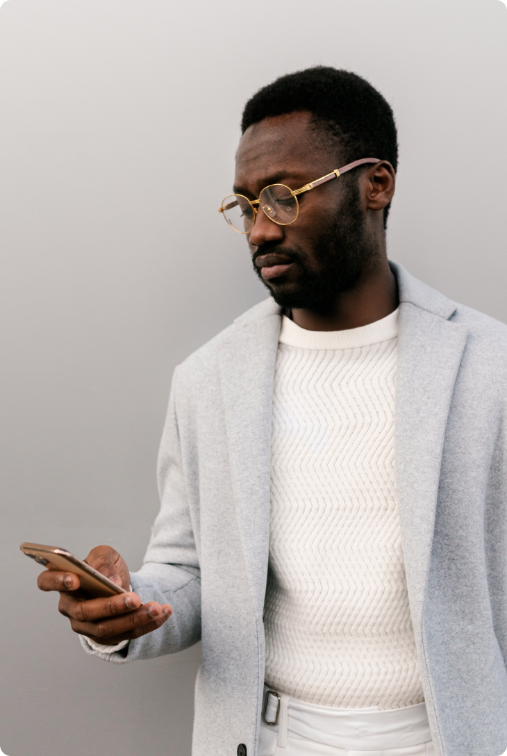 Homem negro de casaco social cinza, com óculos de armação dourada, segurando um celular em sua mão direita.