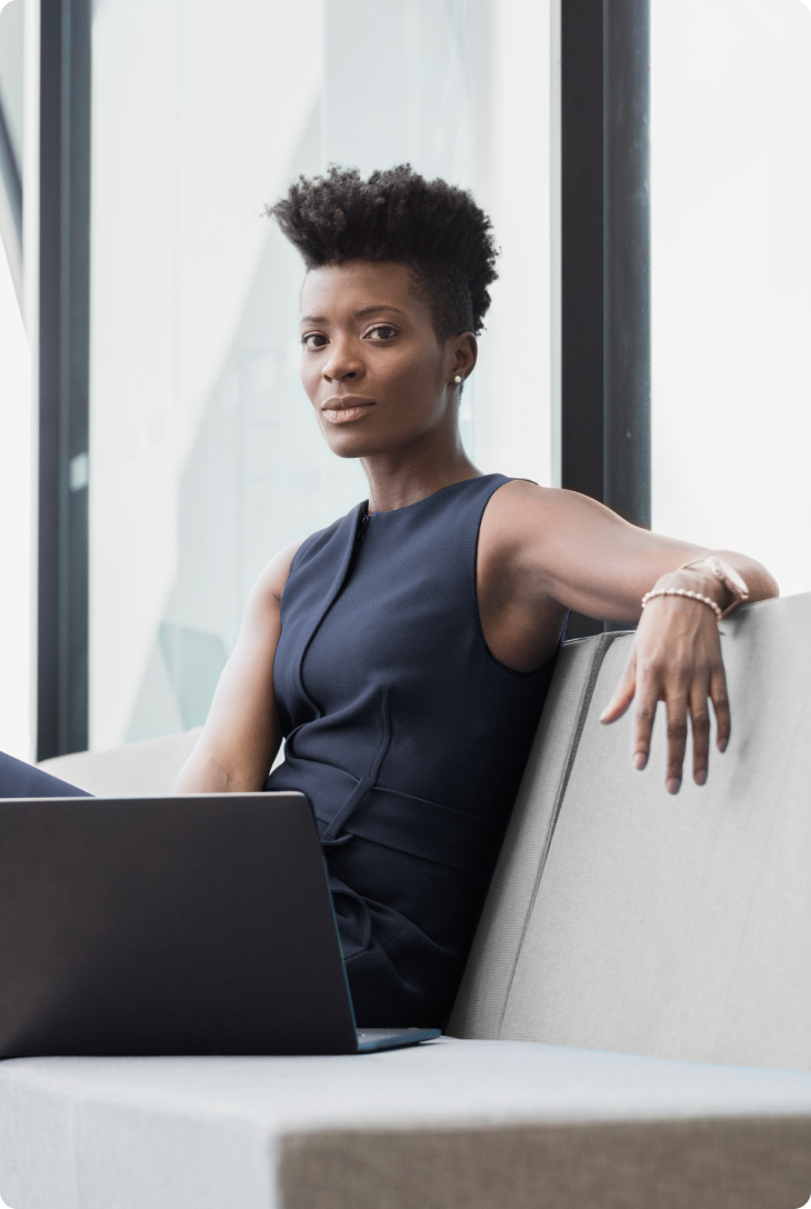 Fotografia de mulher negra, sentada em um sofá cinza, com vestido azul marinho e computador a sua frente