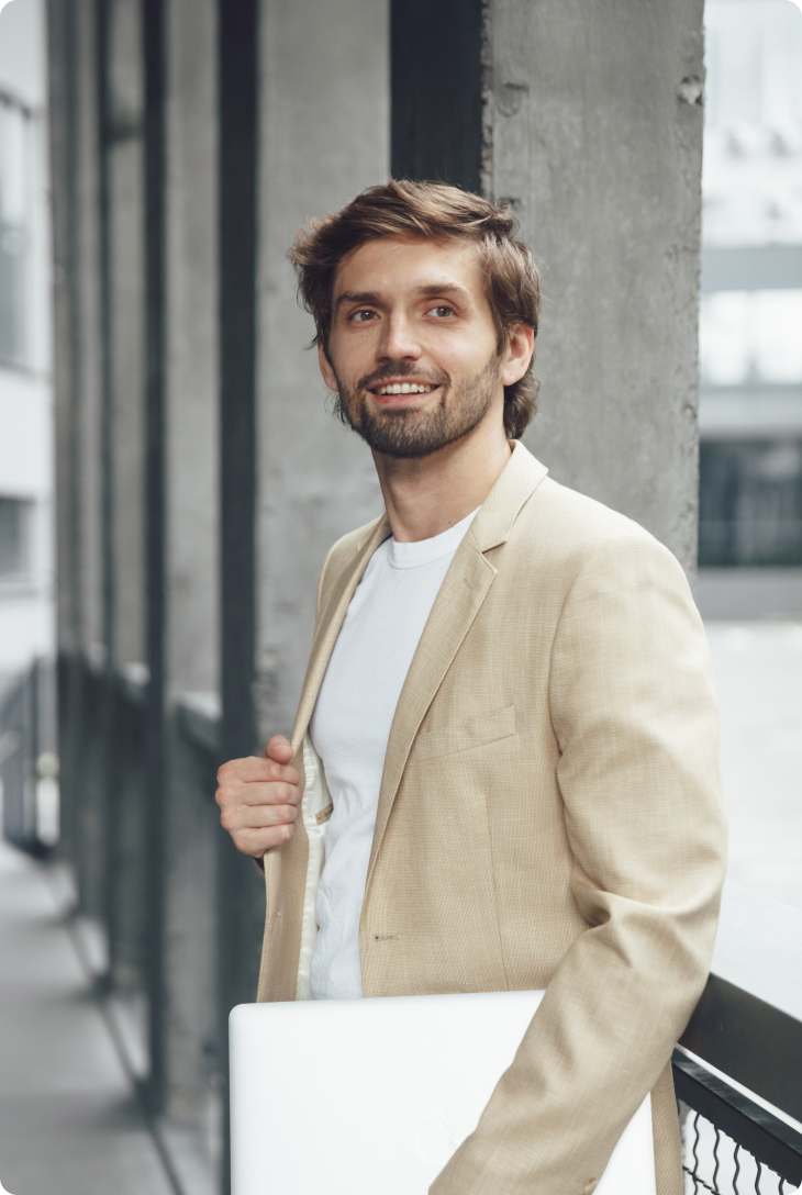 Fotografia de homem com cabelo castanho, de blazer bege e camiseta branca, segurando um computador e encostado em uma grade preta