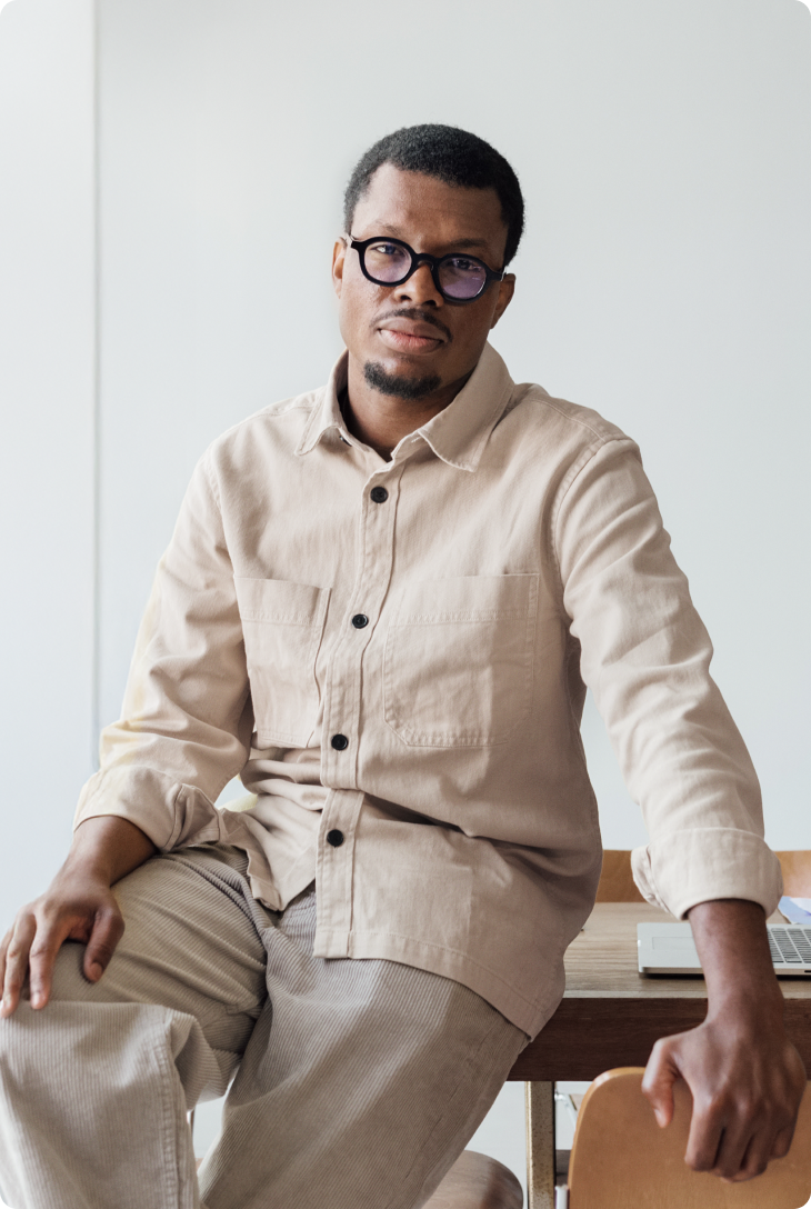 Fotografia de homem negro de camisa e calça bege, usando um óculos de grau preto, apoiado em uma mesa de madeira, com um computador em cima