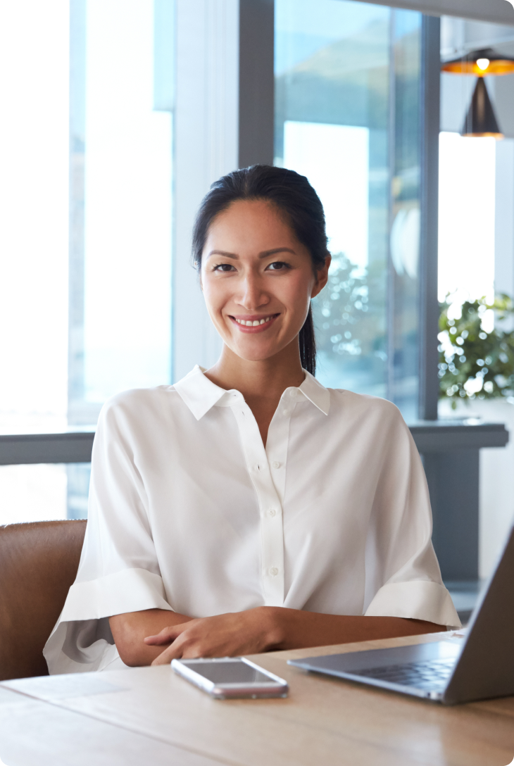 Mulher de camisa branca sentada à mesa com celular e notebook em ambiente corporativo.