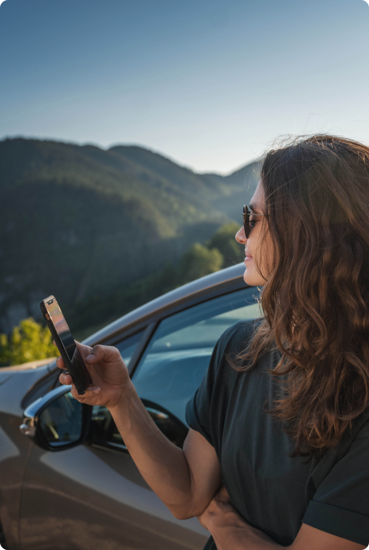 Fotografia de mulher de cabelo cacheado solto, usando óculos escuro, apoiada em um carro, segurando um celular com a mão direita e observando a paisagem de montanhas
