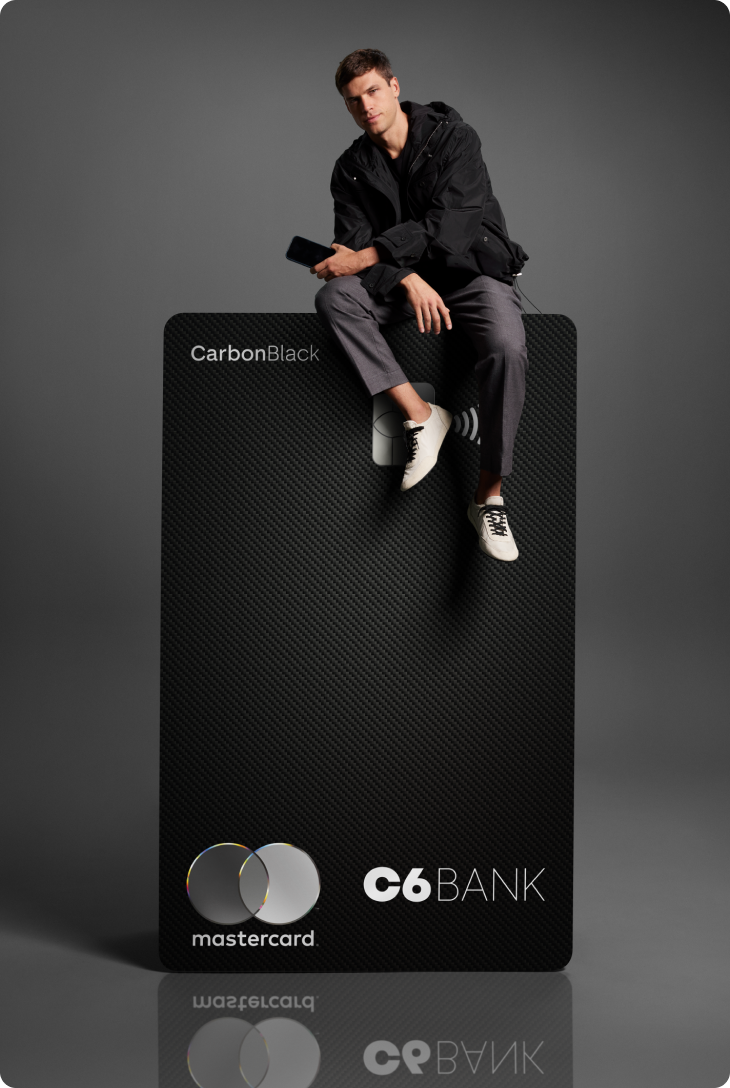 Homem sentado sobre cartão C6 Carbon ampliado, em fundo escuro. Visual elegante e minimalista.