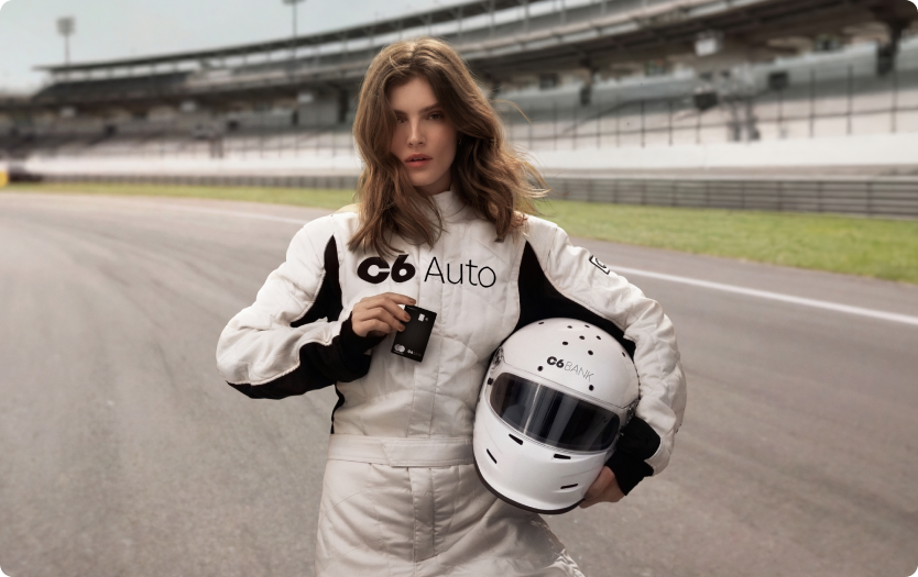 Mulher em pista de corrida com arquibancada ao fundo, usando macacão de automobilismo com o logo C6 Auto, segurando capacete e cartão C6 Carbon na cor preta.
