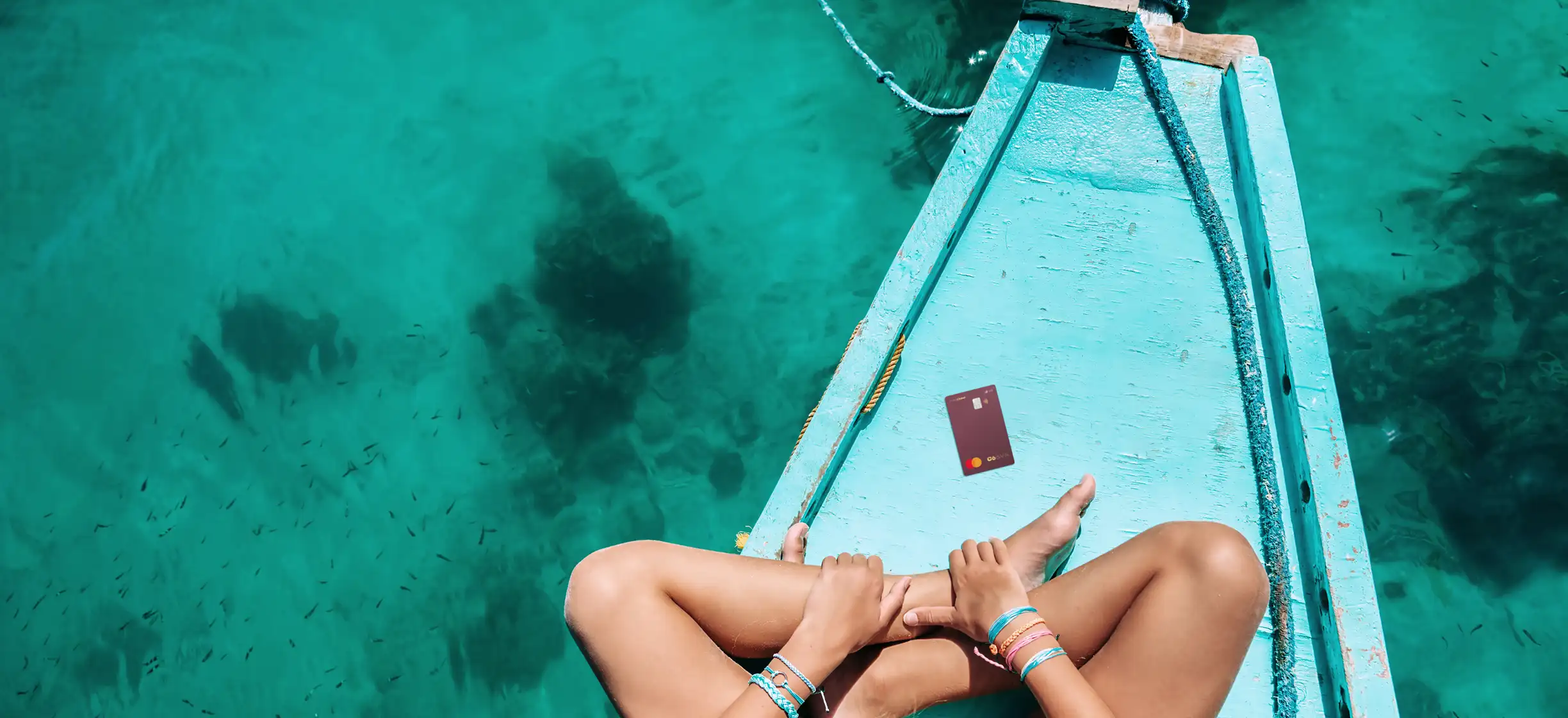 Pessoa sentada na proa de um barco, com o Cartão C6 Conta Global ao lado e o mar cristalino ao fundo.
