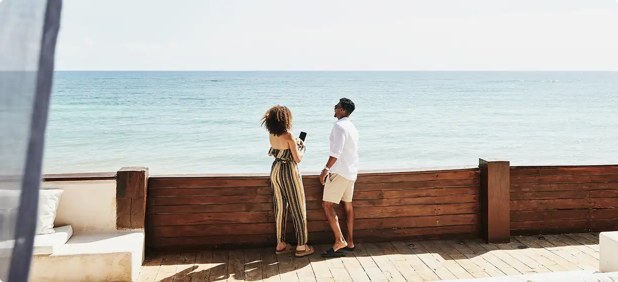 Casal observa o mar de uma varanda com piso e cerca de madeira, em um ambiente sofisticado.
