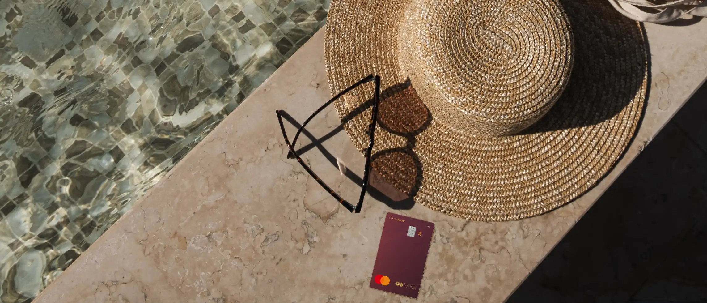 Cartão C6 Conta Global sobre pedra, com chapéu, óculos e piscina ao lado.