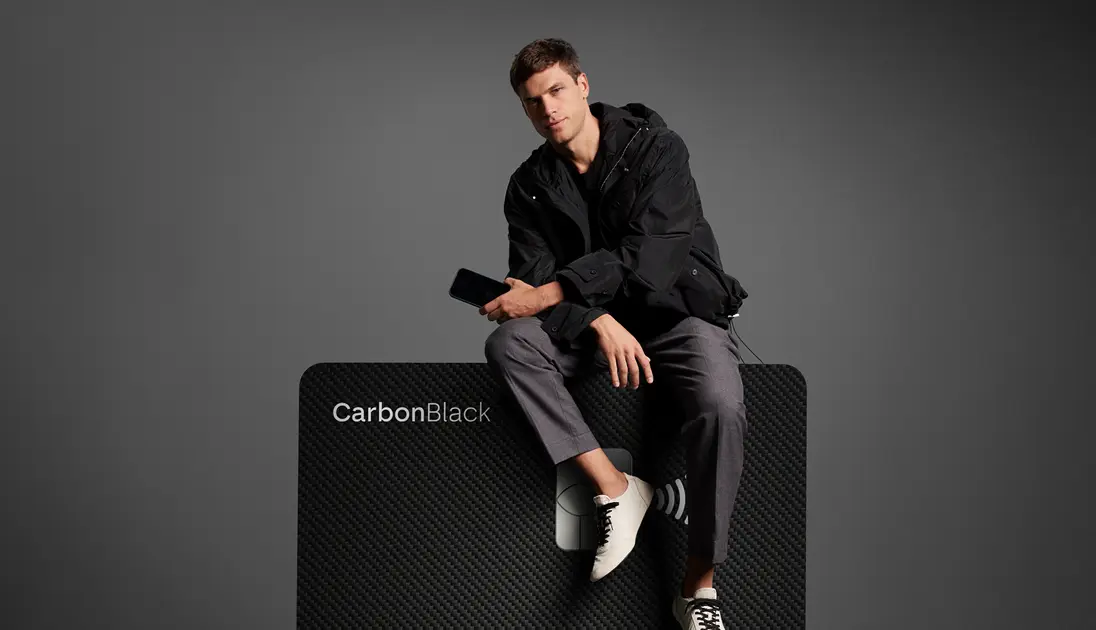 Homem sentado sobre cartão C6 Carbon ampliado, em fundo escuro. Visual elegante e minimalista.