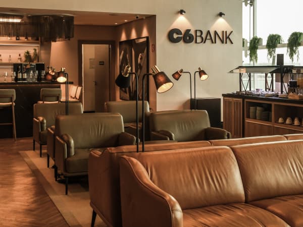 Lounge sofisticado do C6 Bank com poltronas de couro, luminárias e bar ao fundo. A logo do banco aparece na parede iluminada.