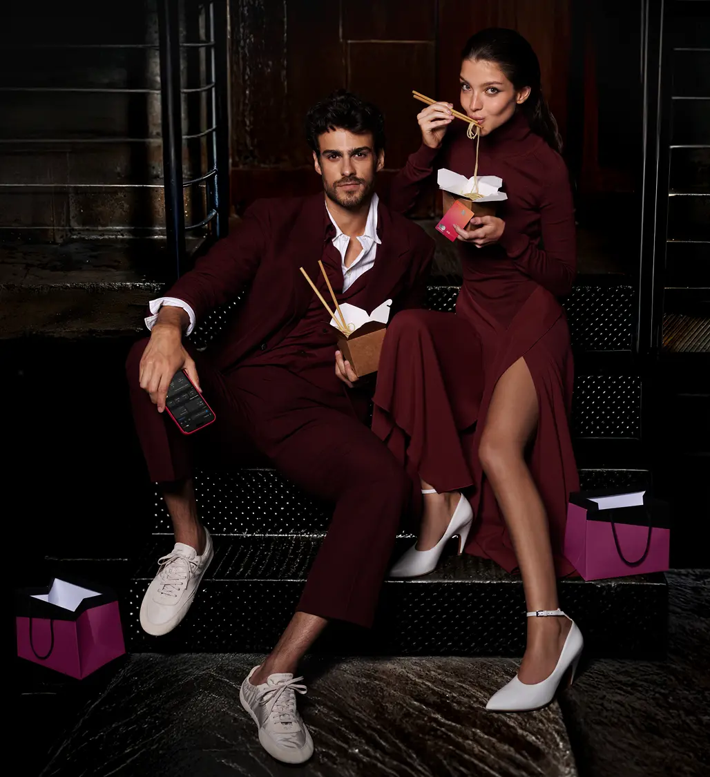 Casal elegante em escadaria com sacolas. Mulher segura Cartão C6 Sunset e o homem, celular com app do C6 Bank.