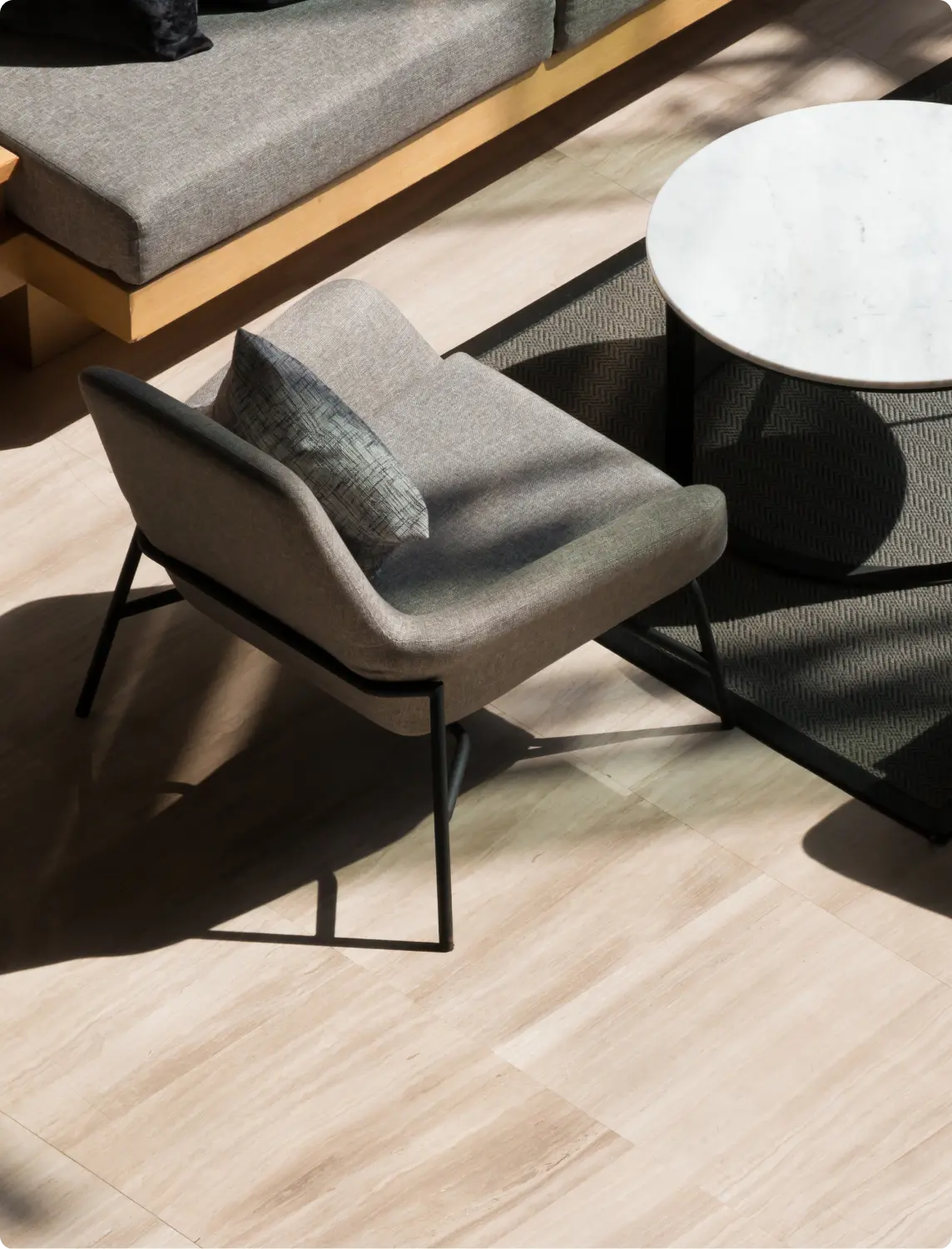 Poltrona cinza com almofada estampada ao lado de uma mesa de mármore redonda, sobre piso claro e tapete geométrico cinza.