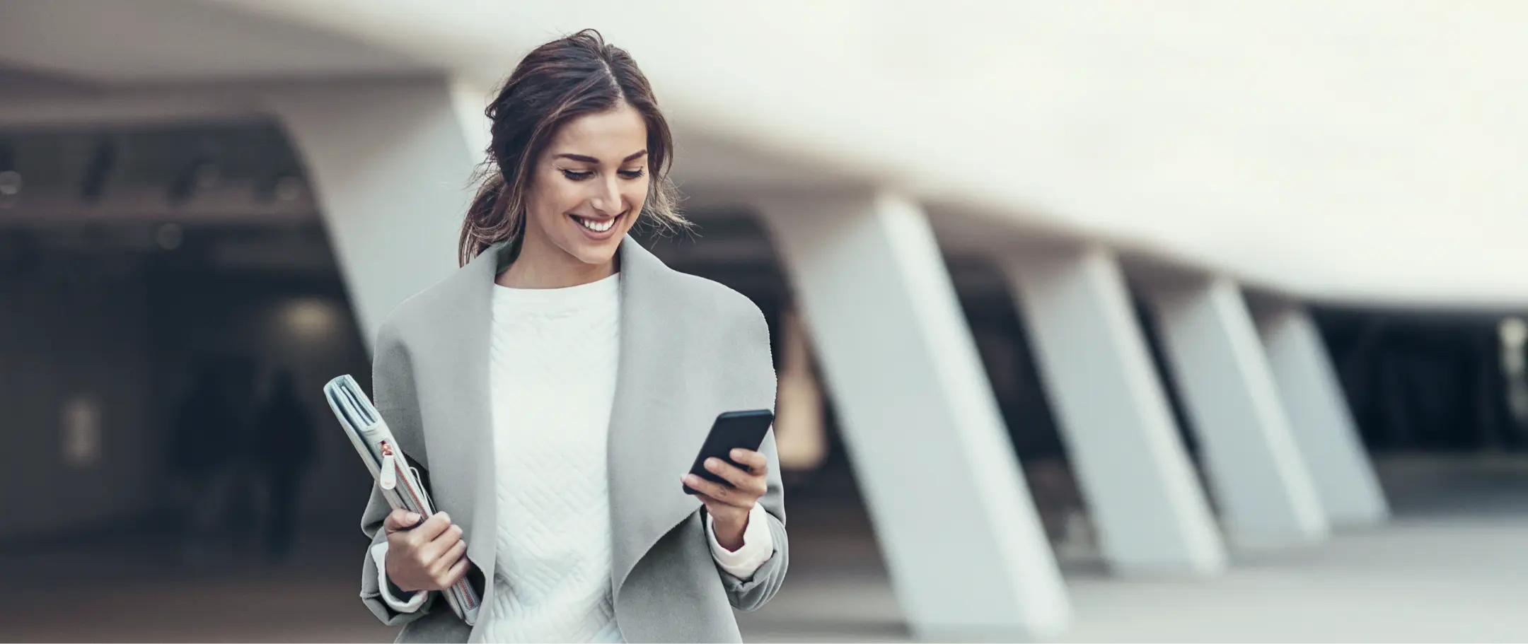 Mulher caminhando sorrindo Mulher sorrindo enquanto caminha com celular e pasta em ambiente urbano.