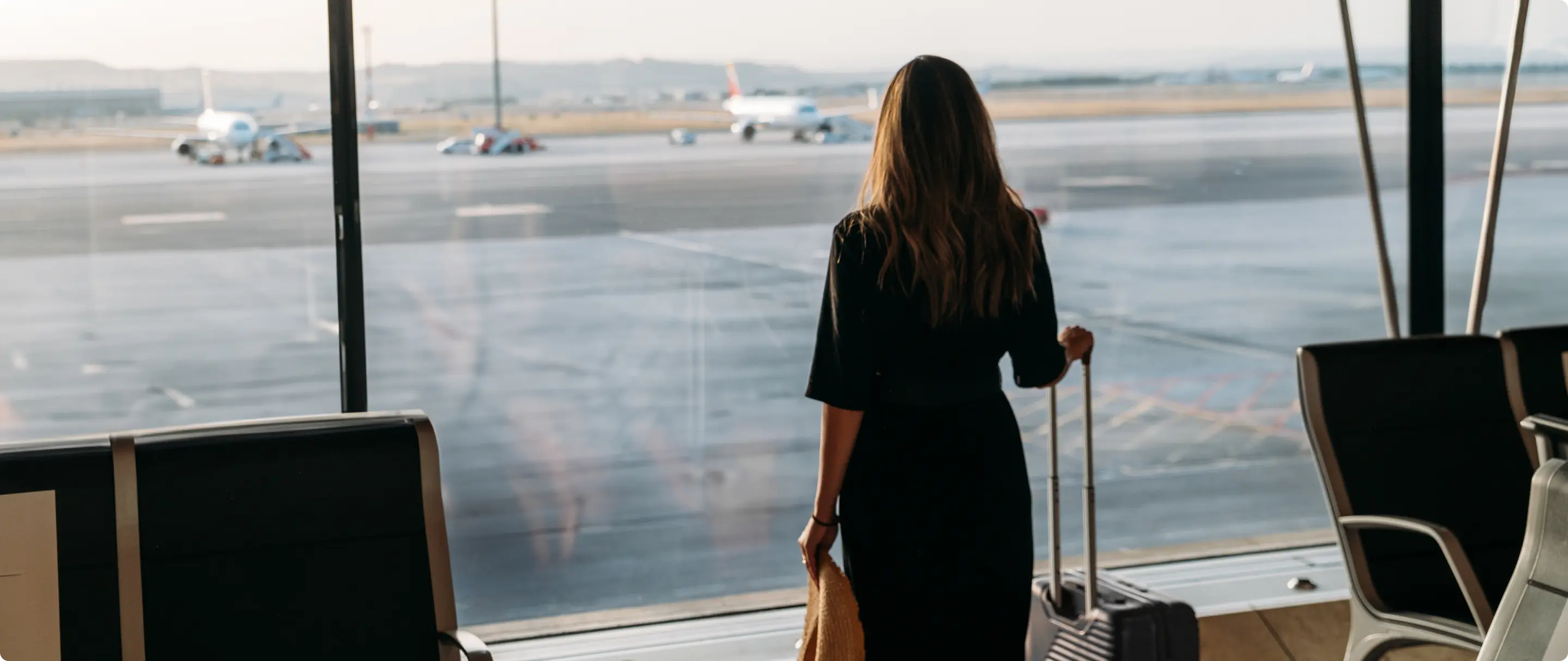 Mulher de costas observa aviões na pista, segurando mala de rodinhas no saguão de um aeroporto.