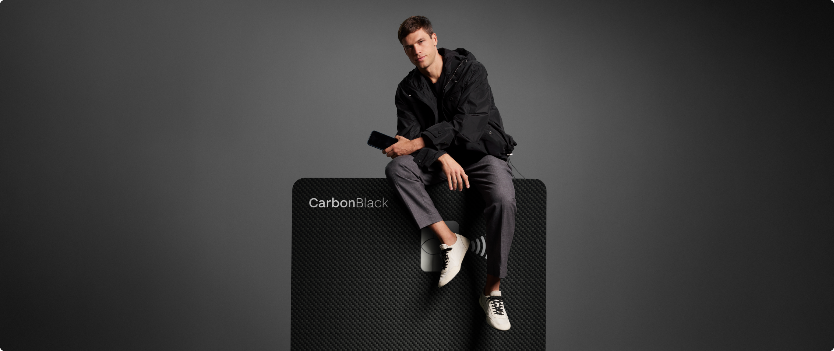 Homem sentado sobre cartão C6 Carbon ampliado, em fundo escuro. Visual elegante e minimalista.