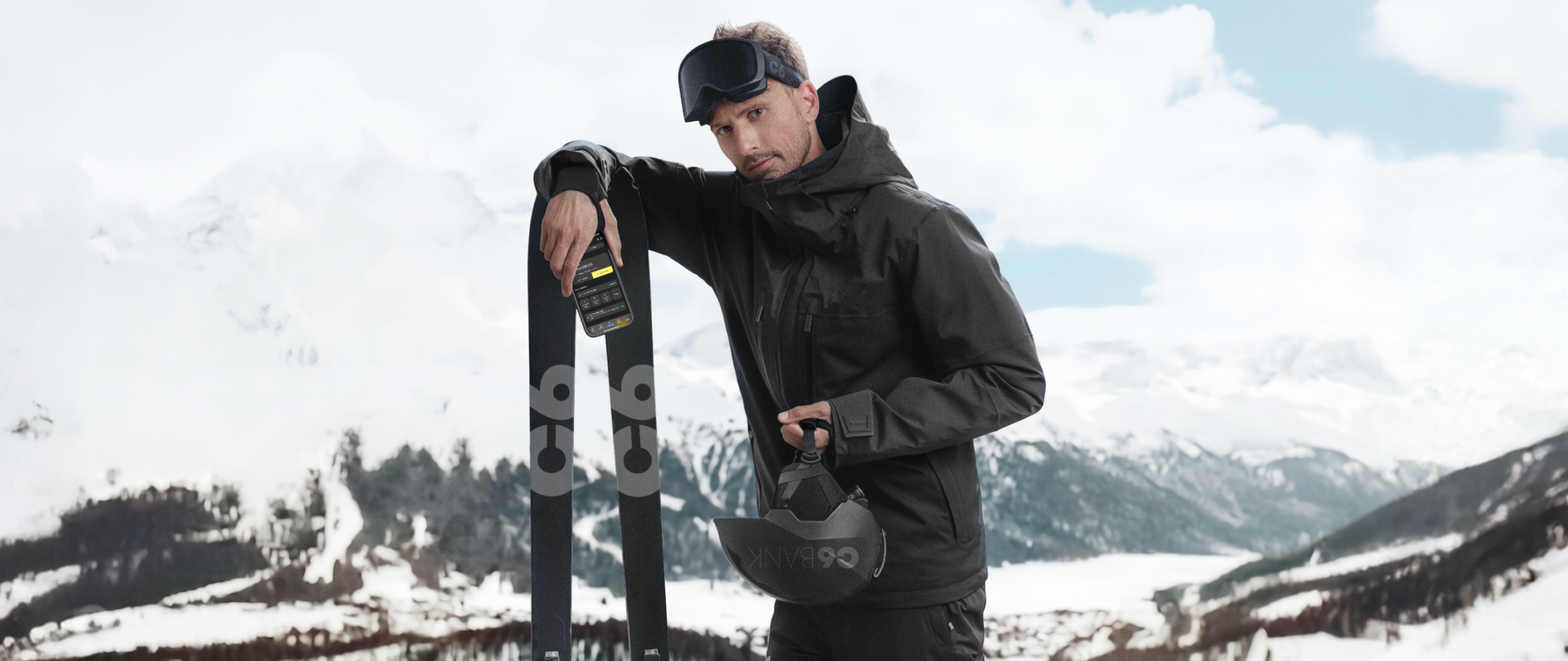 Homem em paisagem de neve com montanhas ao fundo, usando roupa de inverno, apoiada em esquis, segurando capacete, com celular exibindo app do C6 Bank, posicionado sobre o equipamento.